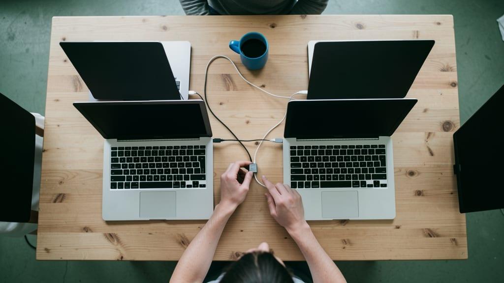 Hands connect a networking cable between two closed laptops on a wooden desk with a blurred blue mug in the background