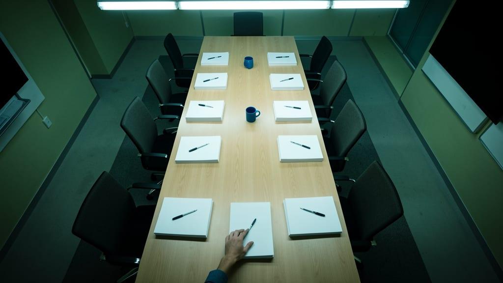 Overhead view of a boardroom table with blank paper stacks and pens at empty chairs as hands slide one stack to the edge